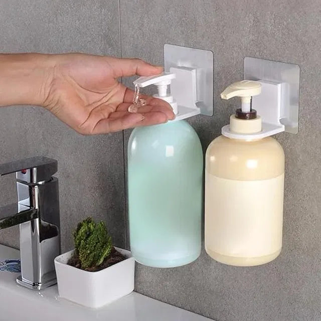 A hand presses the pump of a wall-mounted soap dispenser above a bathroom sink, with the Self Adhesive Shampoo Dispenser Bottle Rack for Bathroom Toilet Kitchen Pack of 2 and a small potted plant nearby.